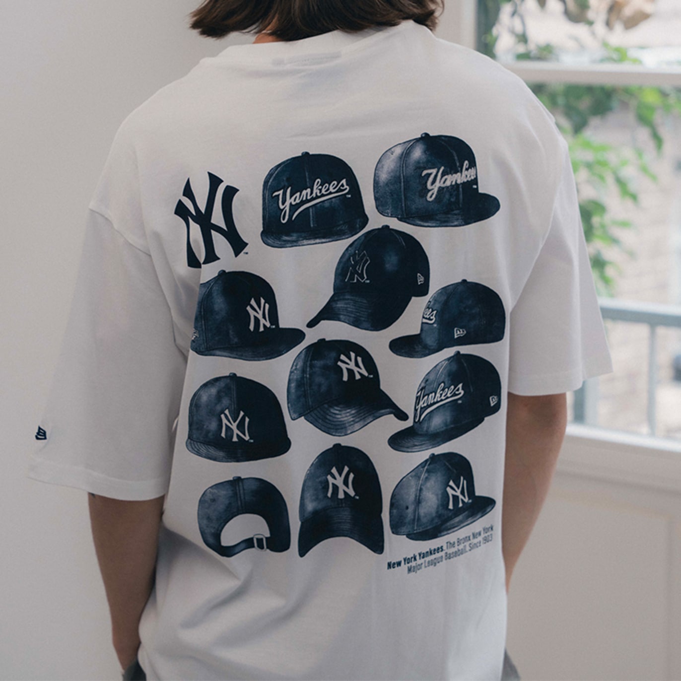Back view of a person wearing a white New Era MLB New York Yankees graphic T-shirt featuring multiple Yankees cap illustrations and the NY logo, standing near a window.