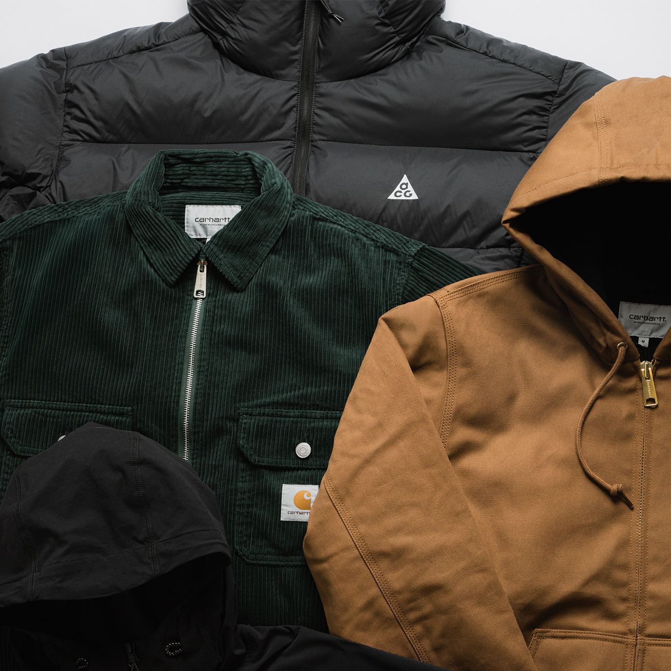 Flat lay of four jackets in different styles and colors: a black puffer jacket with Nike ACG logo, a dark green corduroy Carhartt jacket, a brown Carhartt hooded work jacket, and a black hooded jacket.