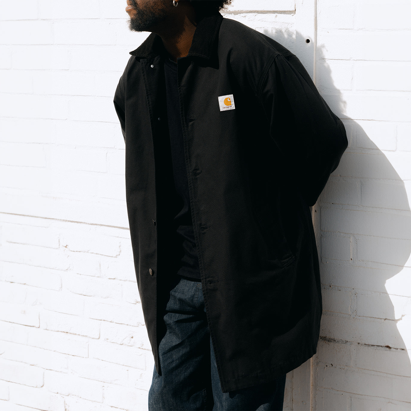 "A man wearing a black Carhartt jacket with a visible logo patch on the chest, standing against a white brick wall in sunlight."