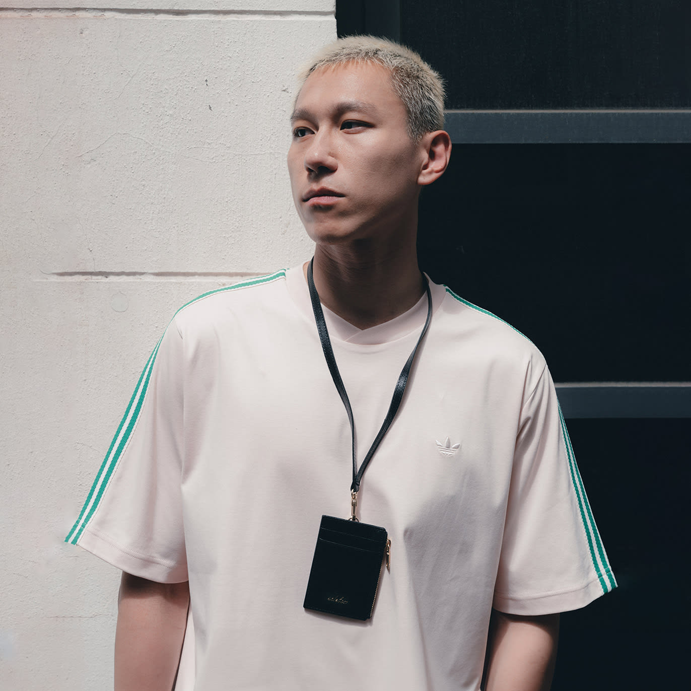 A person with short, light-colored hair is wearing a light pink t-shirt with green stripes on the sleeves and a small white logo on the chest. They have a black lanyard around their neck with a small black pouch attached. The person is looking off to the side, and the background appears to be an exterior wall with a window or dark opening.