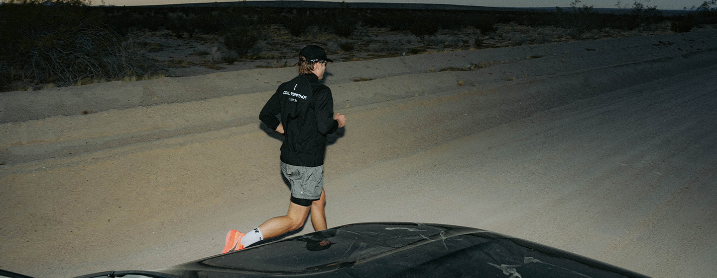 “A man wearing a black jacket, grey shorts, and orange running shoes is running along a desert road at night. The scene is dimly lit by car headlights, and dry desert vegetation lines the roadside.”