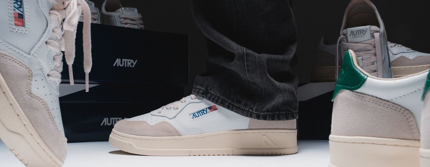 Autry Medalist sneakers in white leather with suede overlays and green heel accents, styled with dark denim and displayed in front of Autry-branded shoeboxes.