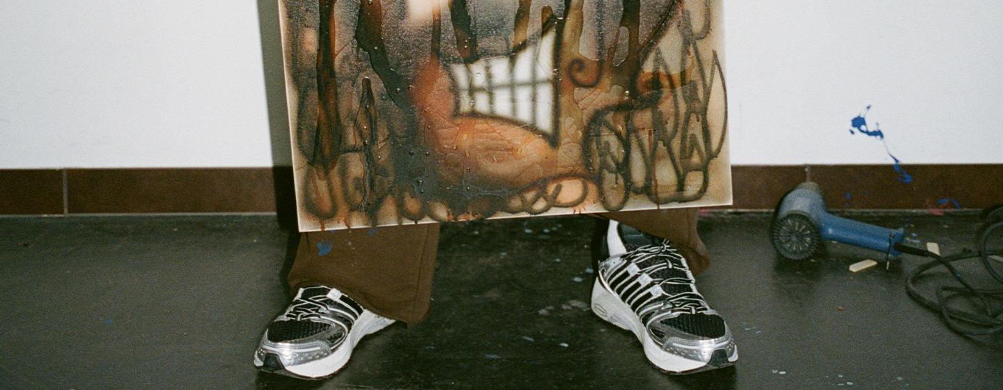 "Person wearing black and white adidas sneakers, standing in a studio with a painted canvas in front of them and tools on the floor."