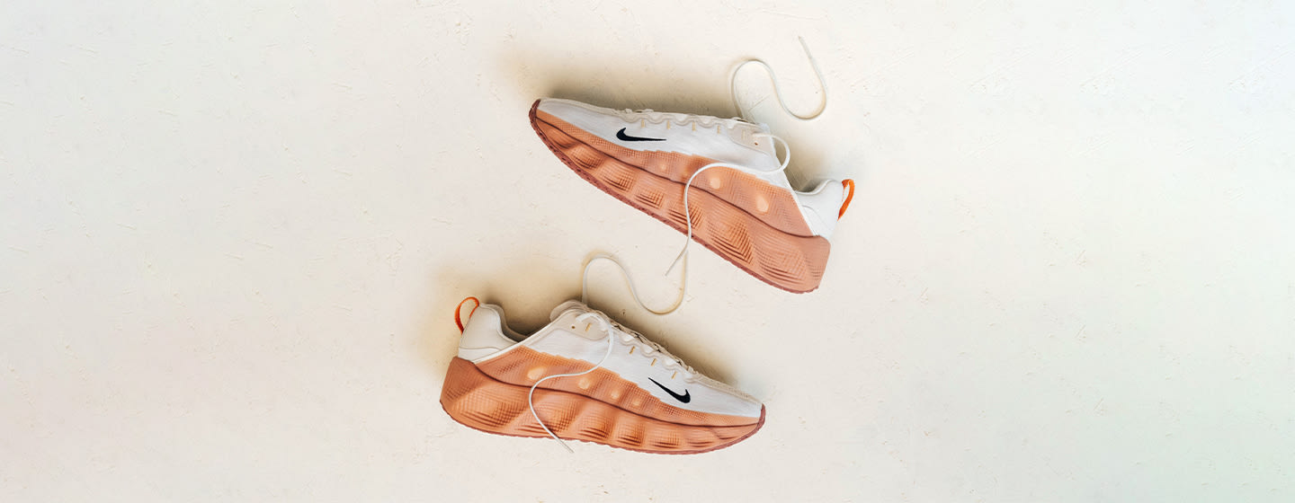 The image features a pair of Nike Ava Rovers, with a white upper and gradient orange sole, set on a clean, neutral surface. The laces are untied, giving a casual, modern look.