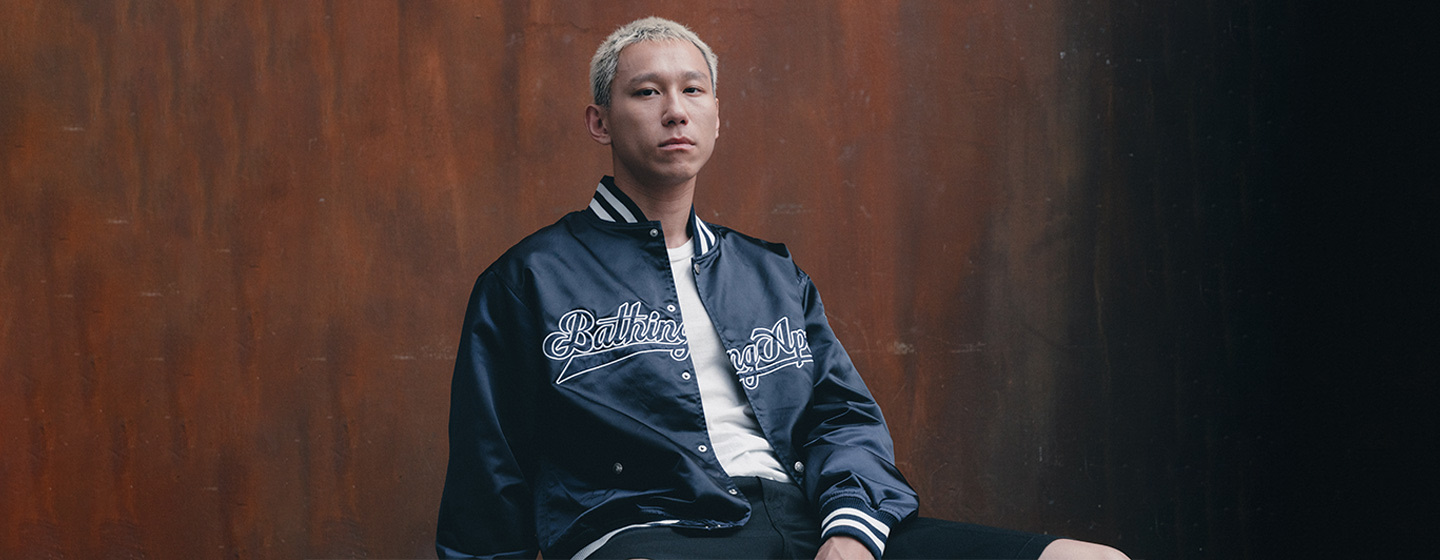 Person with short blonde hair sitting on a black chair, wearing a black satin varsity jacket with white script, a white T-shirt, black shorts, white socks, and white sneakers, posing against a brown industrial-style background.