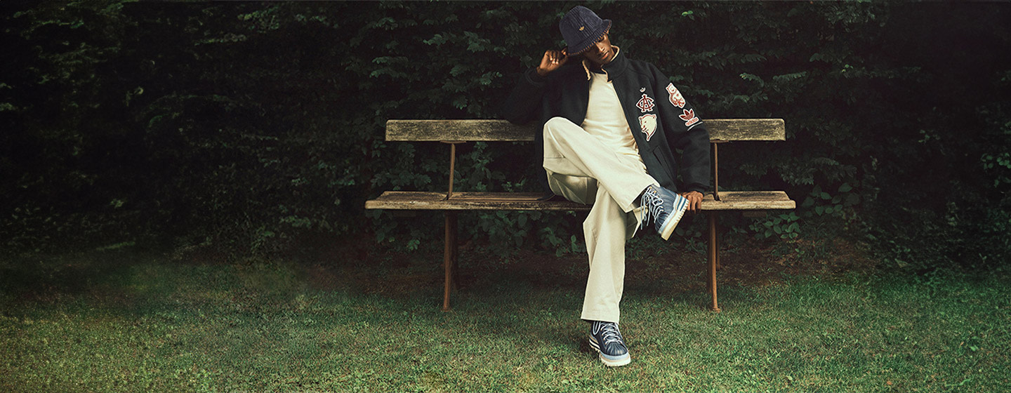Person sitting on a wooden bench outdoors, wearing a black jacket with embroidered patches, light-colored pants, a dark bucket hat, and patterned sneakers, with greenery in the background.
