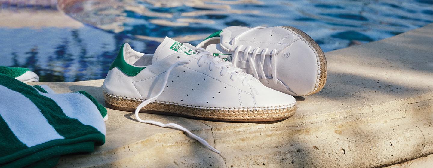 White adidas Stan Smith sneakers with espadrille-style woven soles resting poolside, paired with a green and white striped towel — capturing a relaxed summer vibe.