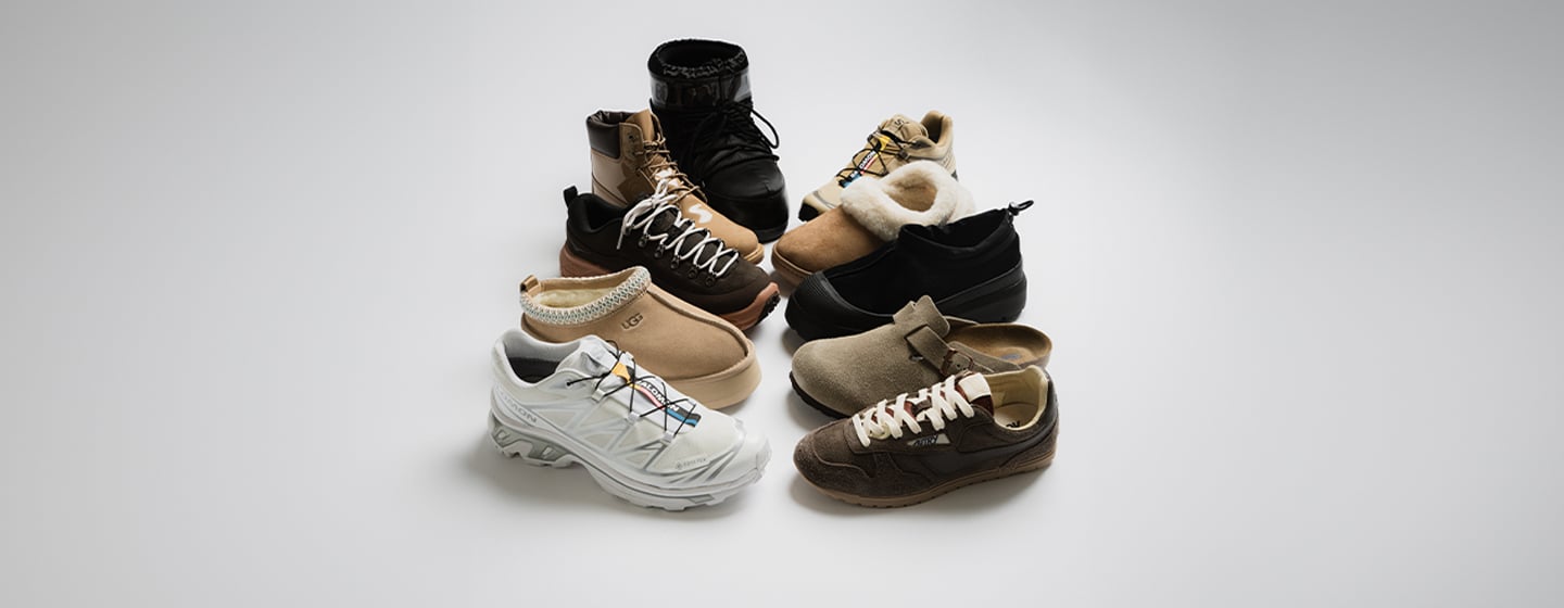 A group of seven different shoes arranged in a semi-circle on a white background. The collection includes a white technical running shoe with colorful lace details, tan moccasin-style shoes with decorative stitching, beige slip-on shoes with shearling lining, black ankle boots with a glossy finish, light beige sneakers with shearling trim, black low-top shoes, and dark brown suede sneakers with white laces.