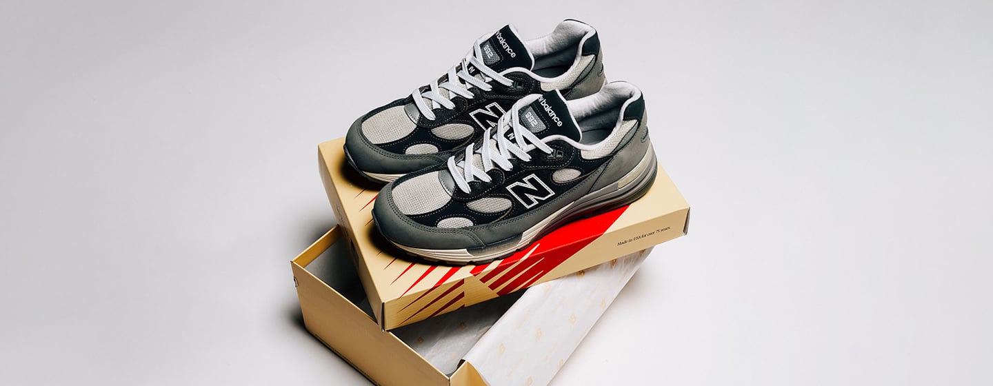 New Balance 992 “Made in USA” (U992GY) grey sneakers placed on top of a beige New Balance shoebox, with the box lid partially open underneath. The shoes feature a combination of grey suede and mesh panels, white laces, and a black “N” logo on the sides. The background is clean and light grey, creating a minimal studio setting.