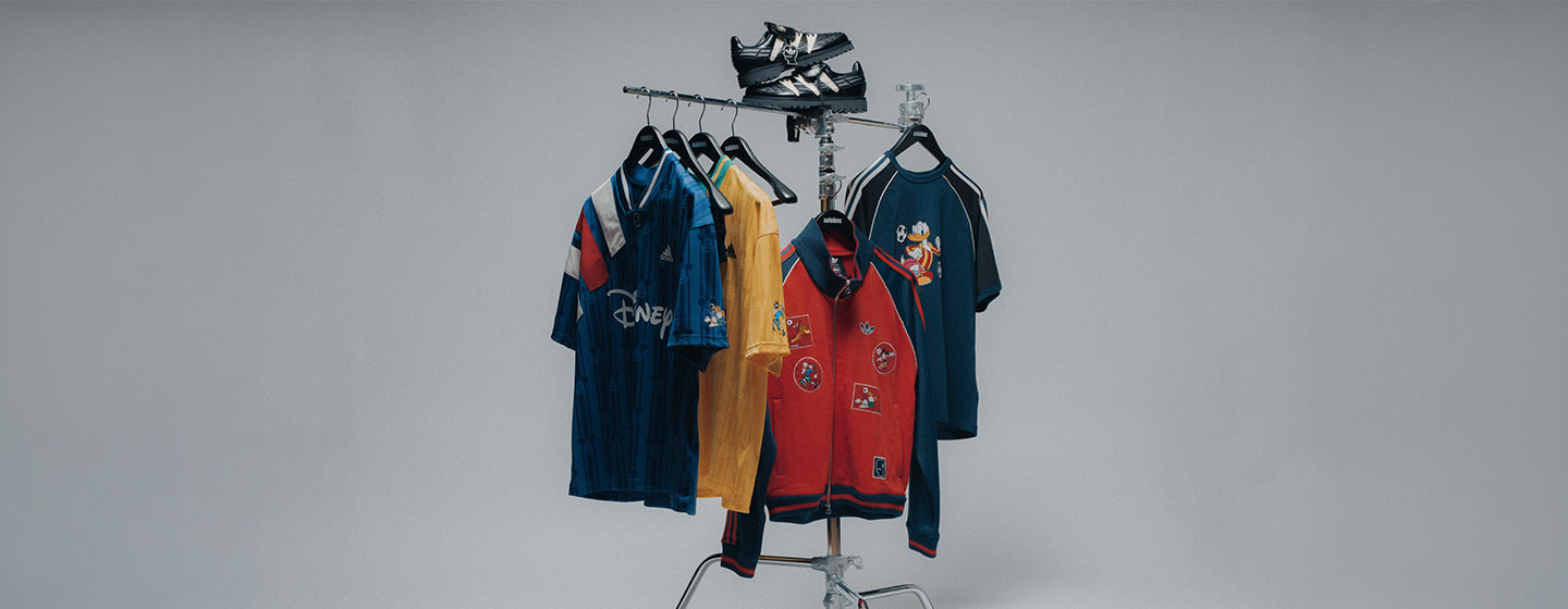 Clothing rack displaying adidas Originals x Brain Dead x Disney apparel, including a red track top with character patches, graphic T-shirts, and jerseys, with matching sneakers placed on top, set against a clean studio background.