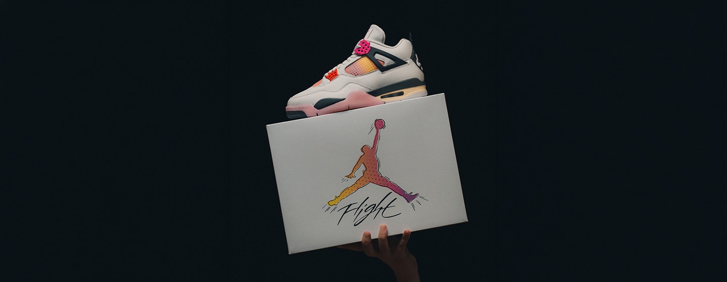 Air Jordan 4 Retro “Comic” sneaker in white, pink, orange, and black displayed on top of a Jordan Flight shoebox, held against a dark background, highlighting the comic-inspired color accents and iconic silhouette.