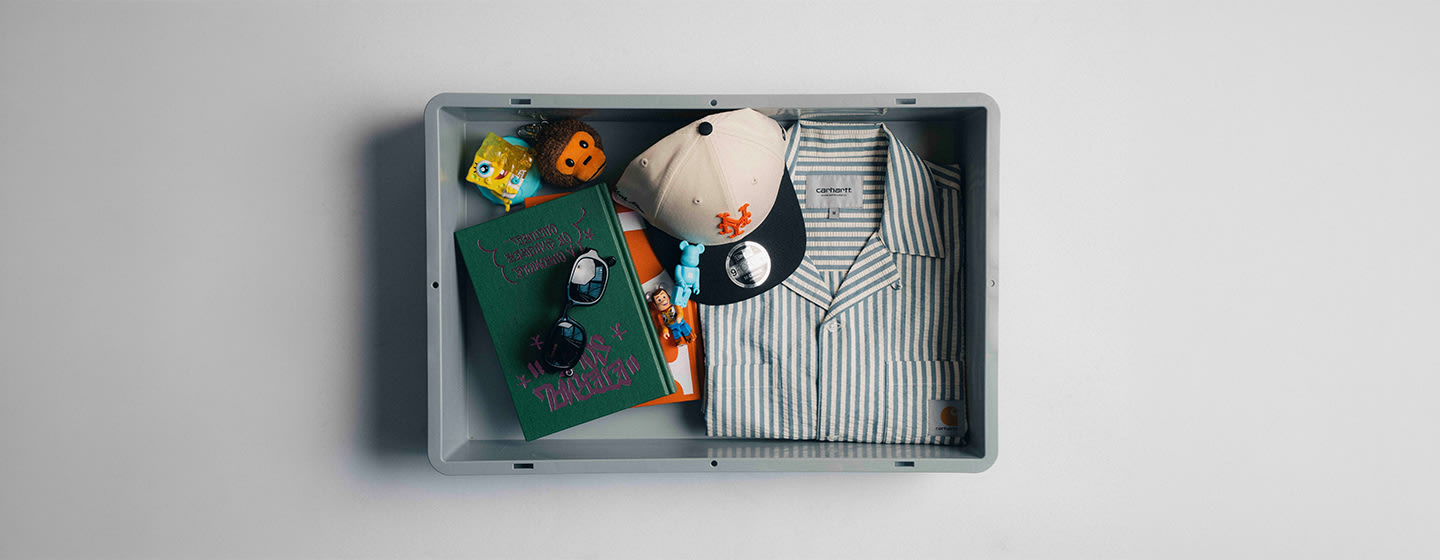 Flat lay of summer essentials arranged in a tray, featuring a striped short-sleeve shirt, beige and black cap, sunglasses, green book, and colorful toy accessories.