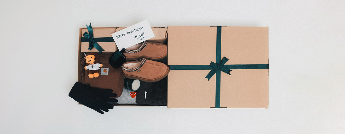 Open cardboard gift box containing assorted items: a pair of tan slippers, black gloves, a brown Carhartt bag, a gray hoodie, a plush dark brown blanket, a small plush lion toy wearing a white shirt, a green mug, and a wrapped present with a green ribbon. A handwritten card inside reads ‘Merry Christmas! Your Toolbox Team.