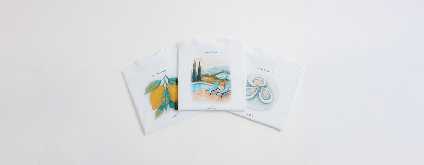 Three folded white T-shirts laid out side by side, each featuring a colorful illustration: lemons on the left, a poolside scene with chairs and umbrella in the center, and oysters on the right.