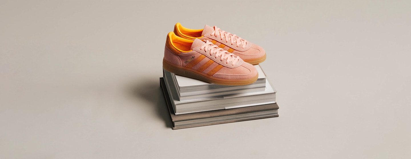 A pair of Adidas Handball Spezial sneakers in soft pink suede with bright orange stripes and inner lining, placed on top of a stack of books against a neutral beige background.