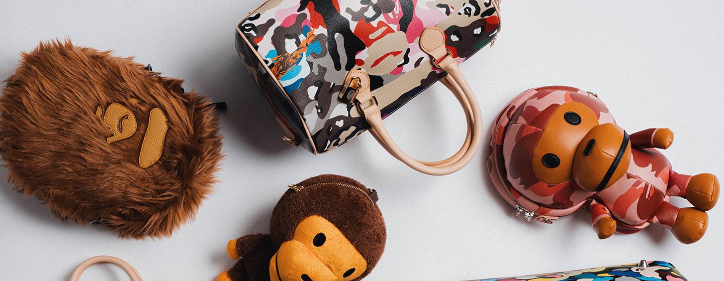Selection of BAPE bags and accessories featuring signature camouflage and colorful ABC Camo prints, displayed on a light grey background alongside BABY MILO character pouches.