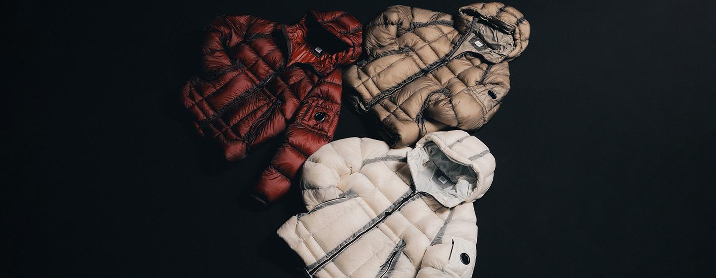 Three C.P. Company puffer jackets in red, beige, and off-white colorways displayed on a dark background, featuring quilted construction, integrated hoods, and the brand’s signature lens detail on the sleeve.