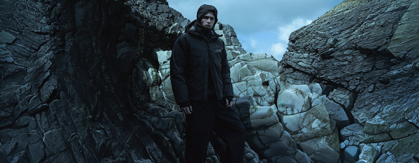 Model wearing a black Purple Mountain Observatory technical jacket and trousers while standing among rugged coastal rocks in an outdoor landscape.