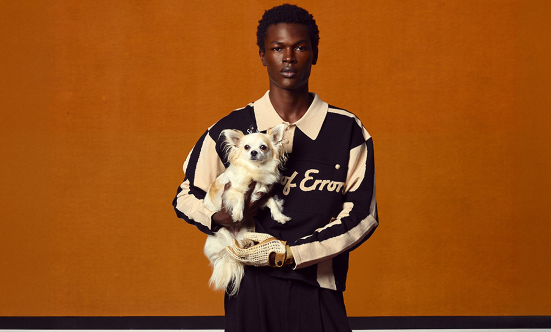 House of Errors campaign featuring a model in a black and cream rugby shirt holding a small white dog against a warm orange backdrop.