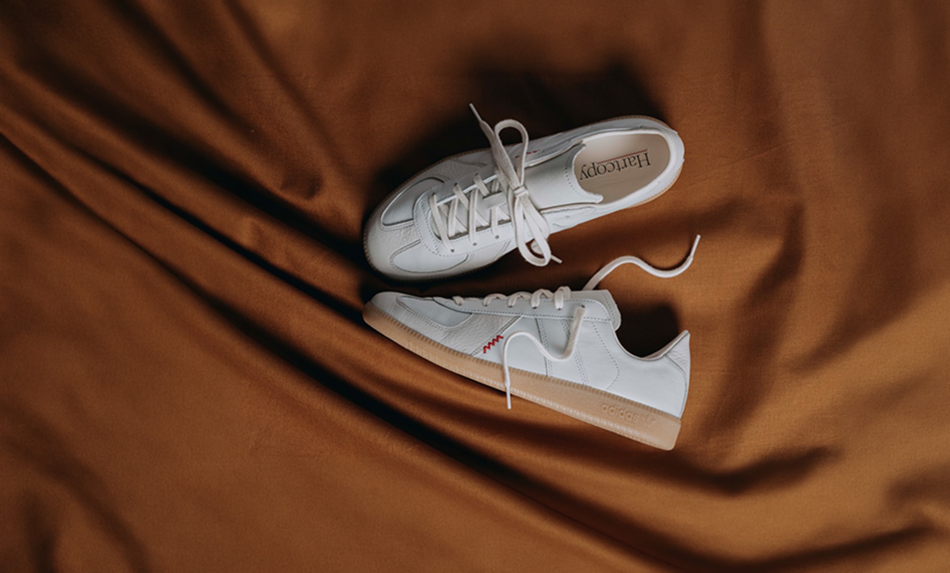 Pair of white adidas sneakers with gum soles displayed on draped brown satin fabric, photographed from above.