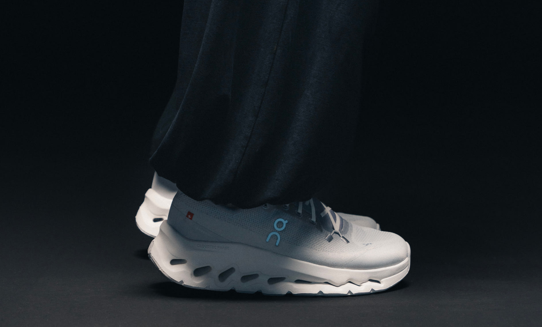 Person wearing white On running shoes with light blue logo details and chunky performance soles, styled with wide black trousers against a dark studio background.
