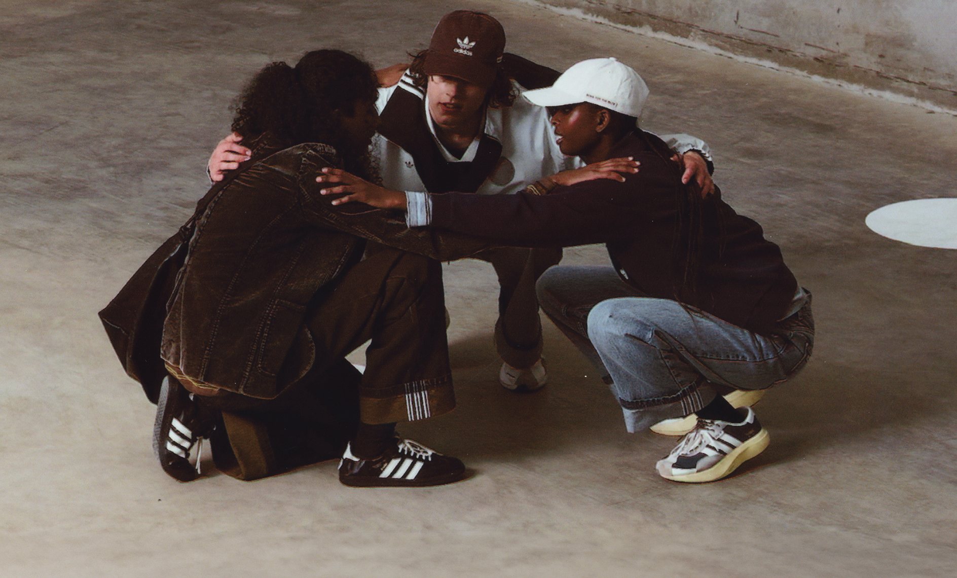 Three models crouching in a circle wearing pieces from the adidas x SONG FOR THE MUTE collection, styled in earthy tones with adidas footwear and apparel in an industrial setting.
