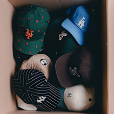 "A cardboard box filled with a variety of baseball caps, featuring different team logos and designs, including a green cap with red roses, a blue LA cap, and several caps with pinstripes and other patterns."