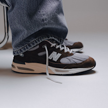 Person wearing brown and grey New Balance 991 sneakers with wide-leg jeans, sitting next to a metal chair on a light grey floor"