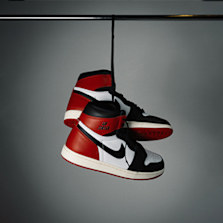 A pair of Air Jordan 1 sneakers in red, black, and white colorway hanging by the laces on a metal bar against a gray background.