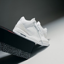 A pair of all-white Air Jordan 3 sneakers leaning against a black shoebox with elephant print details, set against a gradient background.