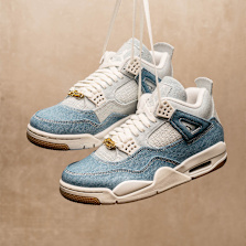 A pair of denim-textured Air Jordan 4 sneakers hanging by their white laces against a beige background. The shoes feature light blue and indigo denim panels, white mesh and leather accents, and gold lace charms.