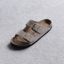 Beige suede Birkenstock Arizona sandals with two adjustable buckles on a textured surface.