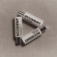 "Three small white bottles labeled 'ACCIDENT' with different scent names: Chlorophyll Gardenia, Celluloid Galbanum, and Radish Vetiver, arranged in a triangle on a textured beige surface."