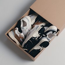 "A cardboard shoebox containing four pairs of stylish sneakers in white, black, and brown tones, neatly arranged on a light grey surface."