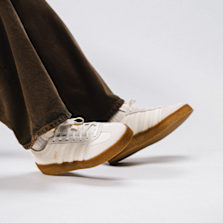 Close-up side view of off-white Adidas Gazelle sneakers with gum soles, worn with dark brown pants and white socks, captured mid-step.