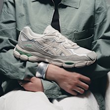 A person in a green jacket holding a light grey and green ASICS GELNYC sneaker close to their chest.