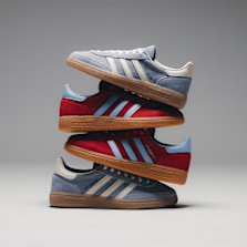 Stack of three Adidas Handball Spezial sneakers in red and blue colorways, each featuring a gum sole and light blue Adidas stripes, against a gray backdrop.