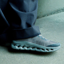 Close-up of the On Cloudtilt sneaker in grey, featuring a lightweight mesh upper and distinctive cushioned sole, worn with dark trousers on a concrete floor.