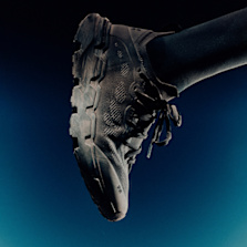 Close-up side view of a black On running shoe with textured upper and rugged sole, worn with a dark sock against a blue gradient background.