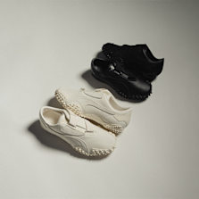 Two pairs of Puma Mostro sneakers in a minimalist studio setting — one in off-white and the other in black, both featuring a sleek slip-on design with distinctive rubber spike soles.