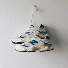 A pair of New Balance 9060 sneakers in beige and navy, hanging by their laces against a plain white wall.