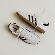 Top-down view of a pair of white Adidas Samba sneakers with dark brown stripes and gum soles, arranged diagonally on a light background.