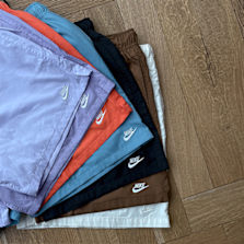 "Stack of colorful Nike athletic shorts in various colors including purple, orange, blue, black, brown, and white, laid out on a wooden floor."