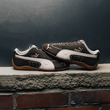 A pair of Puma Speedcat sneakers displayed on a ledge, in a black and white suede colorway with a gum sole, evoking a vintage motorsport aesthetic