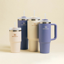 The image features a collection of Stanley brand insulated drinkware, including tumblers and mugs, in soft pastel colors (cream and lavender). The Stanley logo, featuring a winged lion, is prominently displayed on both the left side of the image and on each drinkware item. The background is a simple, warm beige color that complements the products, giving the image a clean and modern aesthetic.