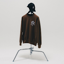 A brown sweatshirt with a large white "NY" style graphic on the chest, displayed on a metal stand with a black cap placed on top.