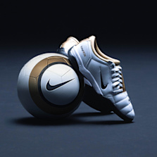 White and gold Nike T90 soccer cleats placed next to a matching Nike soccer ball on a dark surface.