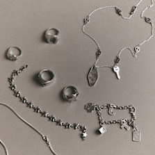 "A collection of silver rings and necklaces laid out on a neutral background, featuring star-shaped designs, keys, a four-leaf clover, a dice, and other small charms."