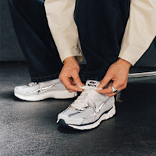 Person tying laces on silver Nike Zoom Vomero 5 running shoes, wearing black pants and a beige jacket.