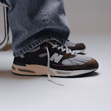 Person wearing brown and grey New Balance 991 sneakers with wide-leg jeans, sitting next to a metal chair on a light grey floor"